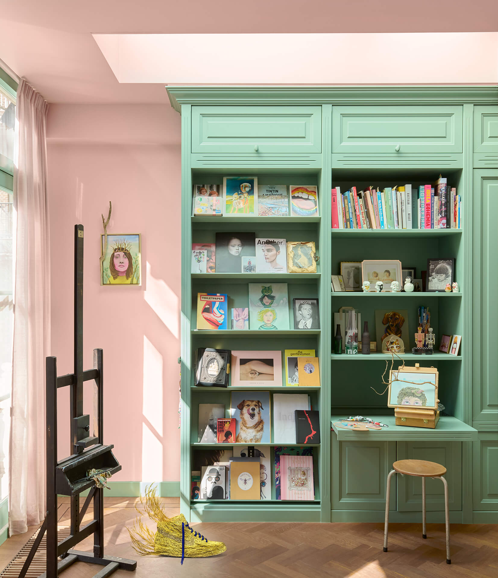 Pink studio with mint green bookcases, an easel and a grass-covered sneaker sculpture