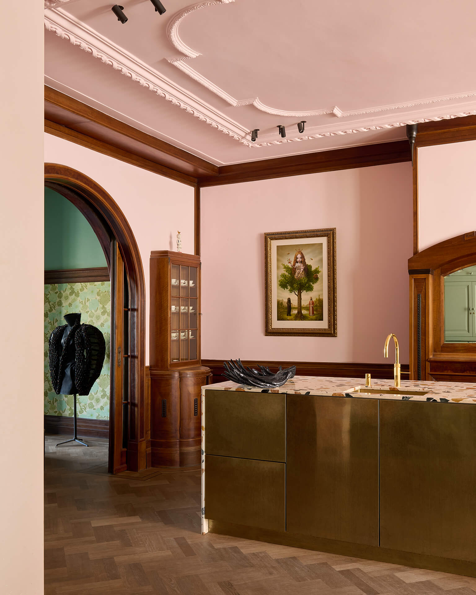 Pink kitchen with brass cabinets, a framed painting and a black couture piece in the doorway