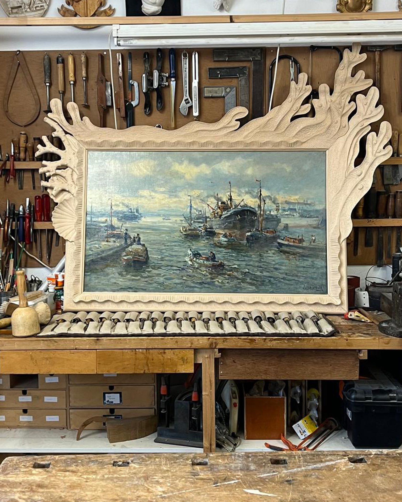 Workshop wall of tools behind a carved wooden coral frame around a harbour painting